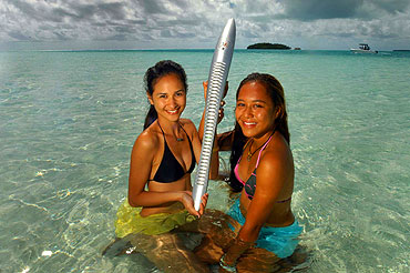 Aitutaki. Eikura and Teau. On a sand bar, Eikura and Teau hold the baton surrounded by crystal blue water.